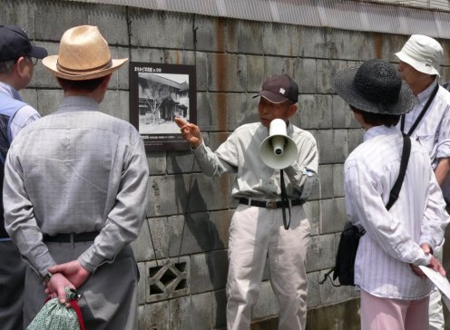 かつて日野町役場があったところにたつ民家の壁に貼られた写真＜昭和26(1951)年 松本保氏撮影＞を解説する松本保さん（北原在住）。現在の中央公民館入口の道路を 挟んだ西側付近です。